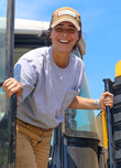 Una trabajadora agrícola desciende de un tractor amarillo mientras sonríe.